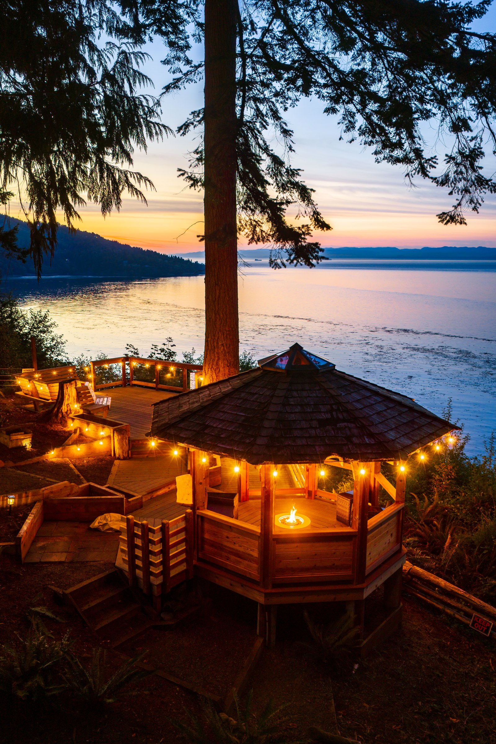Aerial of a string light gazebo and fire pit on a deck above the water at sunset at a Dreamspace Vacations rental near Port Angeles, Washington